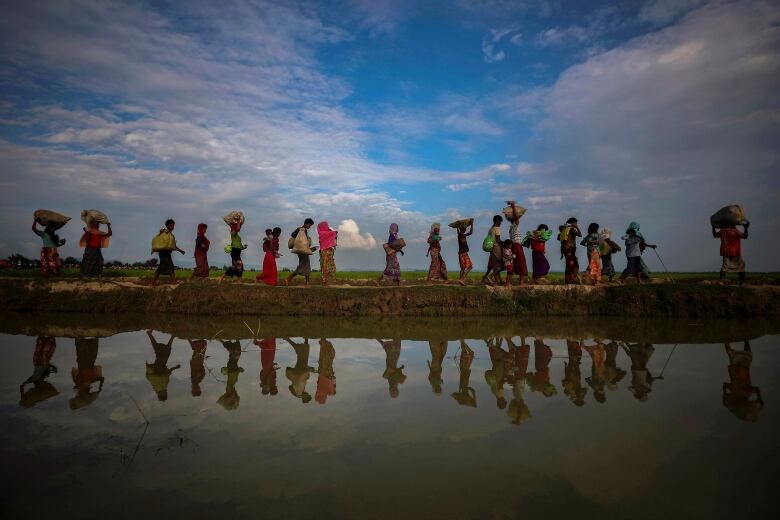 People in a row walk alongside the water holding bundles of their belongings. Their reflections are visible in the water. 