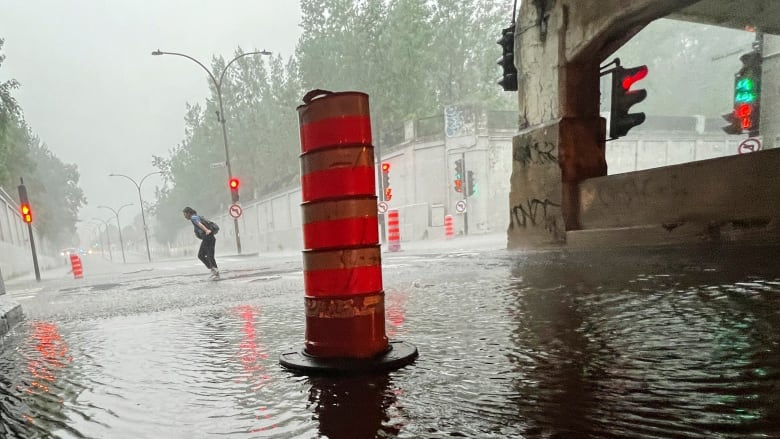 A woman in caught in heavy rainfall as she crosses an intersection with several flooded underpasses. An orange construction cone is placed in the middle of one of them.