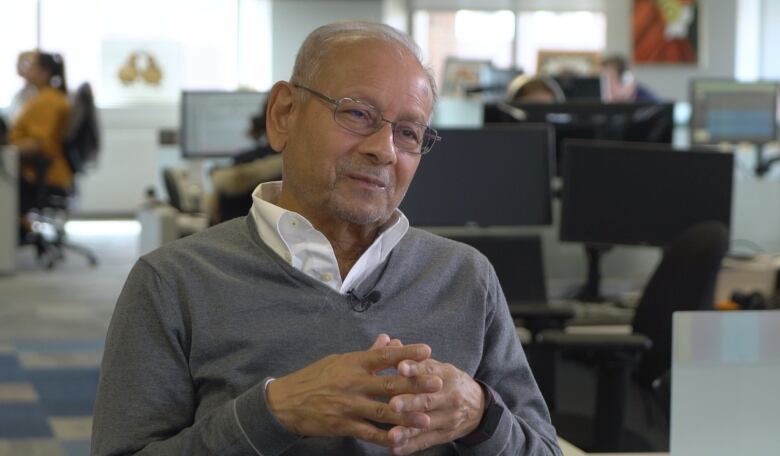 An older man sits in a chair with his fingers intertwined.