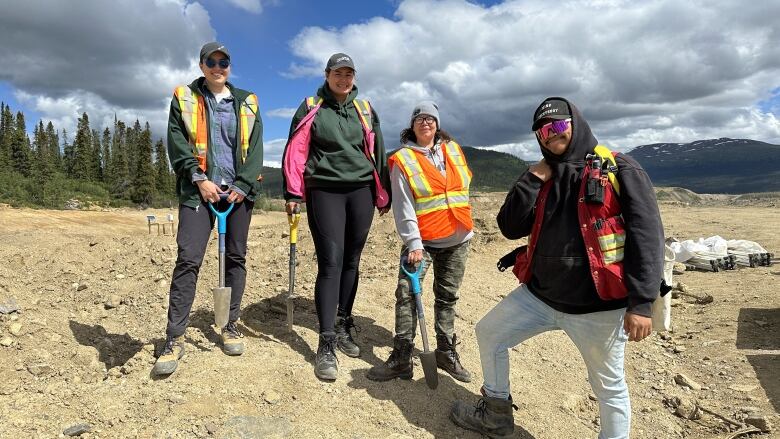 Four people in safety vests holding shovels pose on a rocky field. 