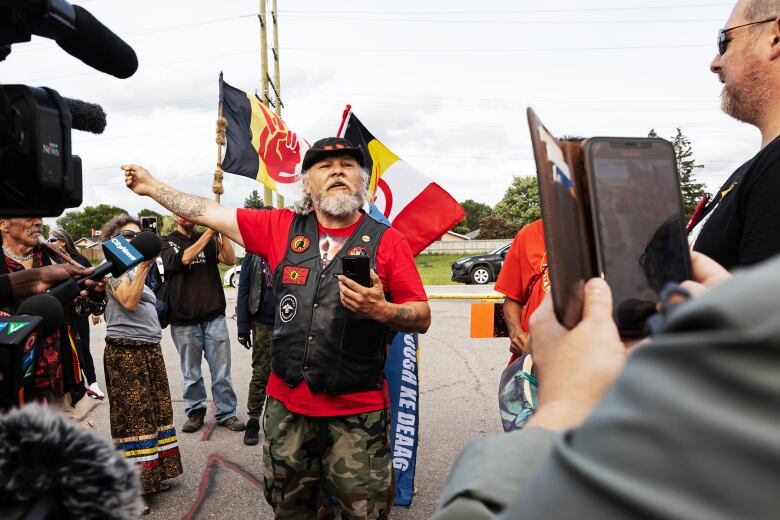 A man holding a cellphone holds his arm out and points to his right. People holding flags stand behind him.