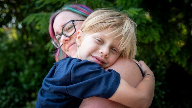 A woman with pink, blue and purple coloured hair hugs a blonde boy who smiles at the camera.