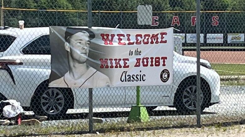 A banner with a grey-scale picture of a man in a baseball uniform with red and black letters saying Welcome to the Mike Buist Classic hangs from a chain link fence. 