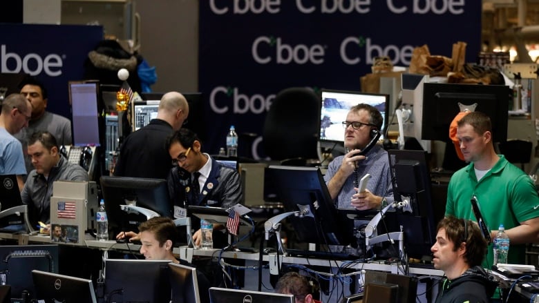 Stock traders in casual clothes sit in front of computers with Cboe branding behind them. 