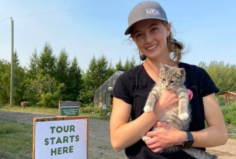 Farmer Charlotte Wasylik, wearing a ball cap and smiling, holds a kitten on her family farm near Vermilion, AB. There is a sign to her left that says 