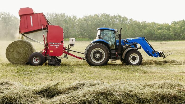 A tractor pulls a hay baler across a field. Piles of hay are seen in the foreground.