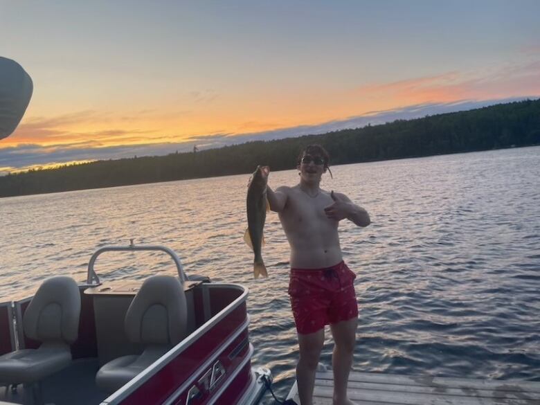 A man in a swimsuit holds up a fish in front of a lake. 