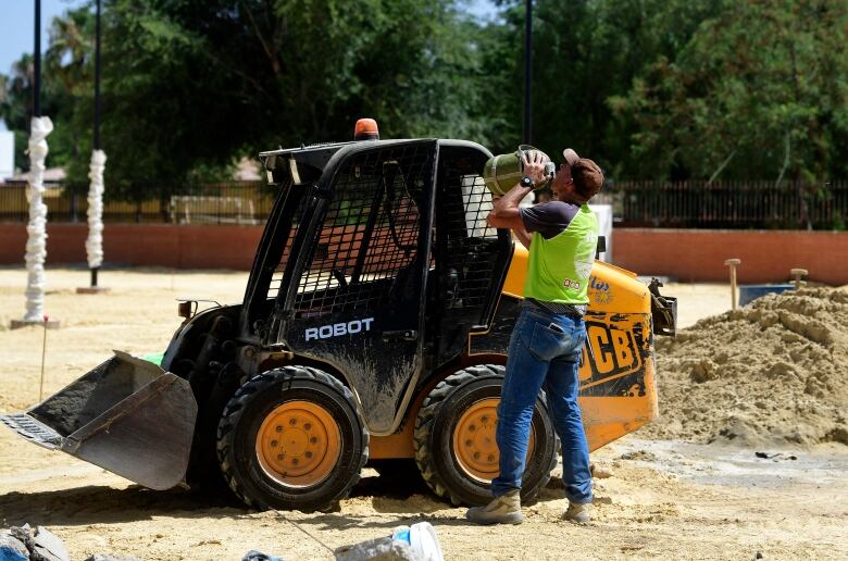 A mean in jeans, workboots and a reflective vest takes a drink from a large jug while standing beside a digger.