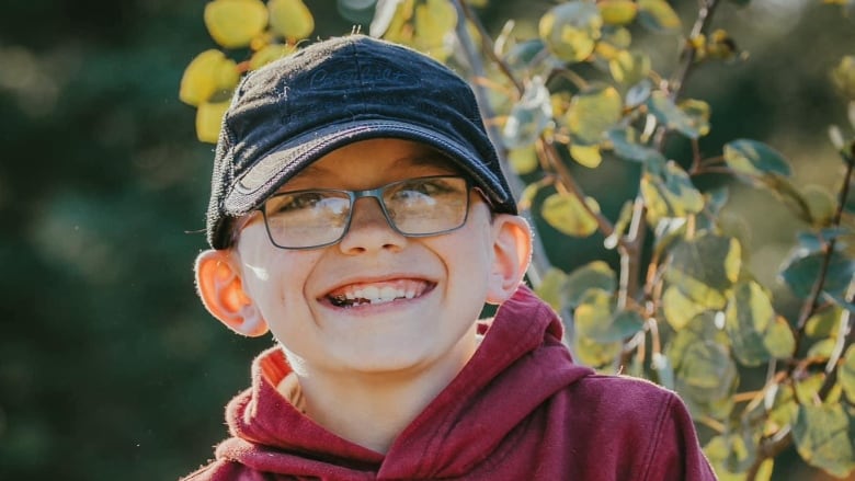 A young boy wears a black baseball cap, blue-rimmed glasses and a burgundy hoodie and smiles for a photo in a park.