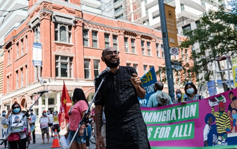 A man in a black shirt with a mic in front of him, surrounded by banners and flags. 