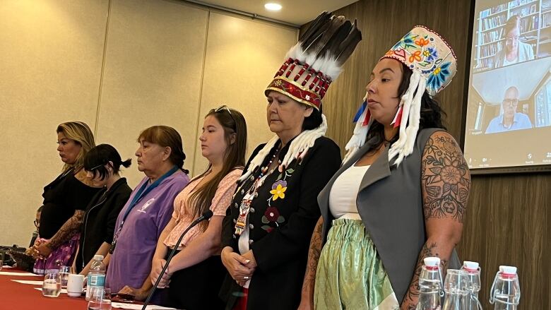 Six women, two in headdresses, stand at a table in front of microphones.