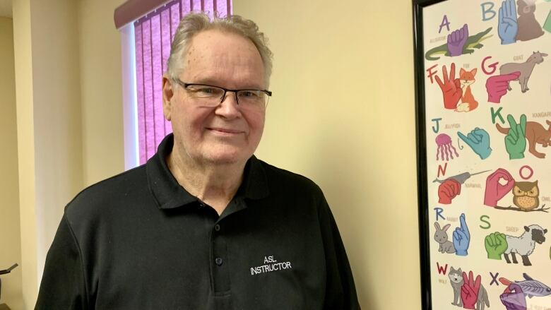 A man wearing a black polo shirt and glasses smiles beside a picture that shows the American Sign Language alphabet.