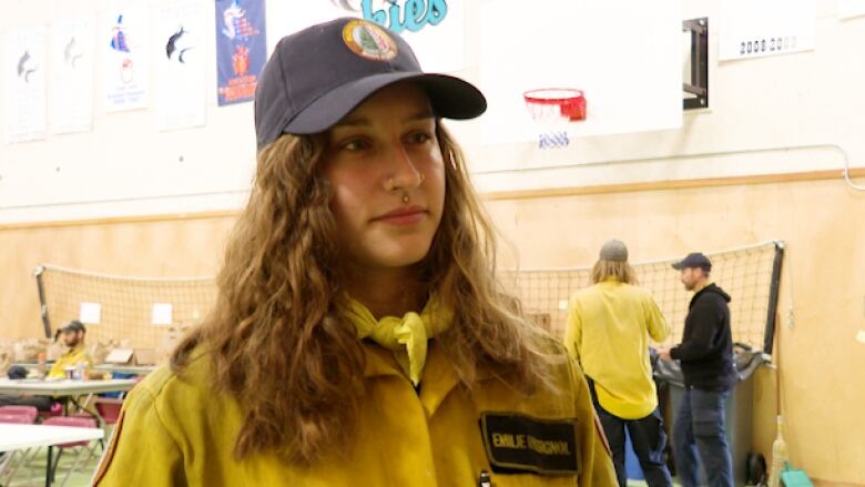 A young woman in a yellow jacket and cap stands in a gymnasium.