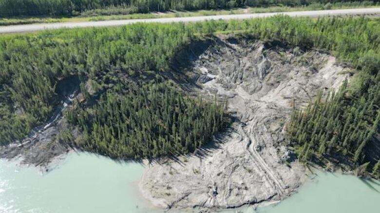 An aerial shot of a large muddy slump seen descending into a river, with a road visible behind the slump.