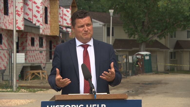 A man in a suit stands behind a podium in front of housing under construction.