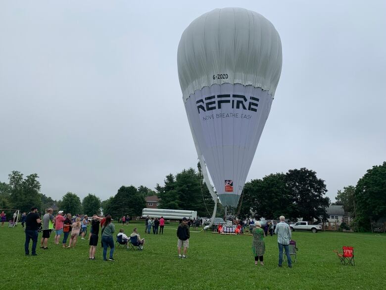 A giant white transatlantic helium balloon stands inflated in a field with many nearby onlookers.