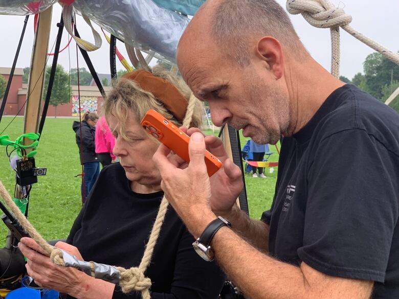 A woman with short blonde hair wearing a black tee-shirt and a man that is nearly bald with a black tee-shirt stand looking at their phones, facing the left of the frame. They are standing in the basket of a hot air balloon, only evident from the ropes nearby.