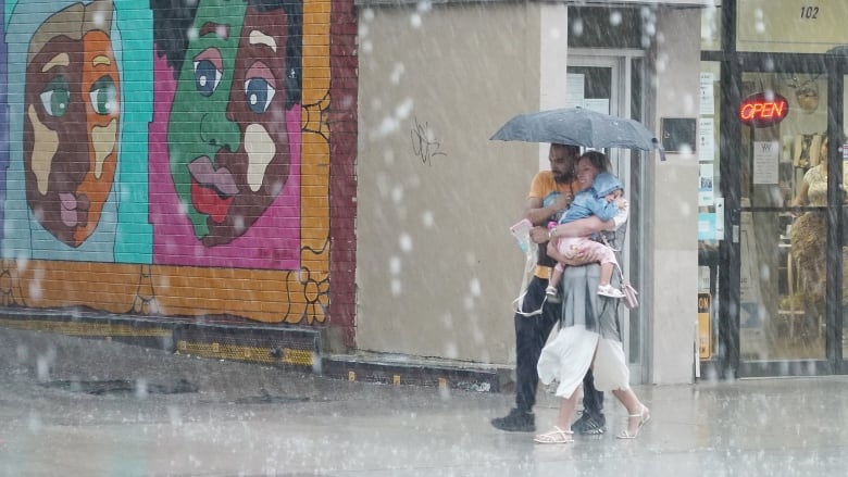 A man and woman - the woman is carrying a child - walk under an umbrella during heavy rain in a downtown core