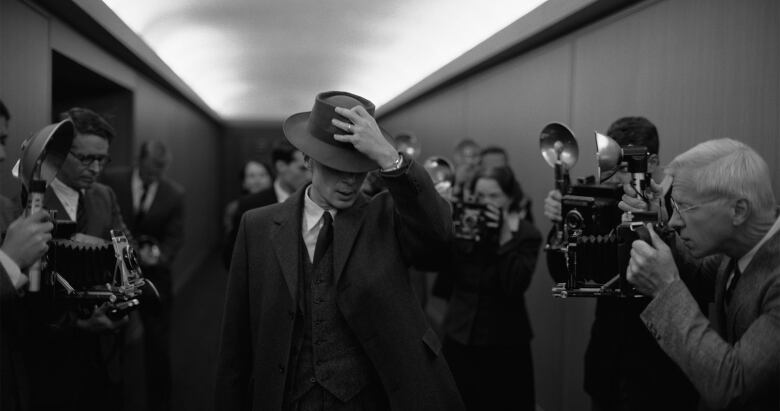 A black and white image of a man with a fedora pulling it down over his face. On either side of him photographers try to take his picture. 