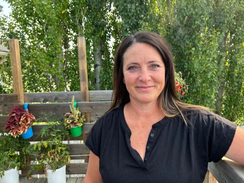 A close-up shot of a woman in a black shirt as she stands on a deck with plants and trees behind her.