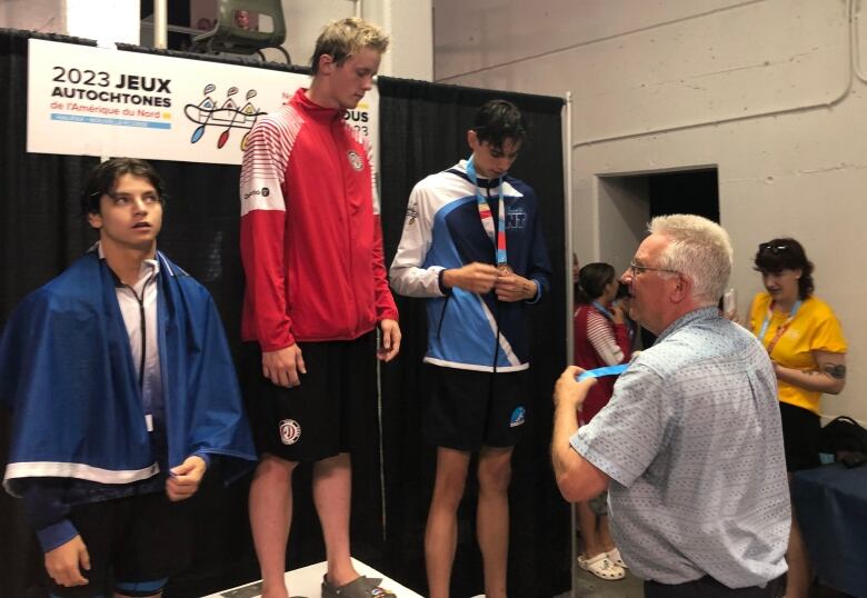 Three young man stand on a medal podium as an older mean holds a medal in front of them.