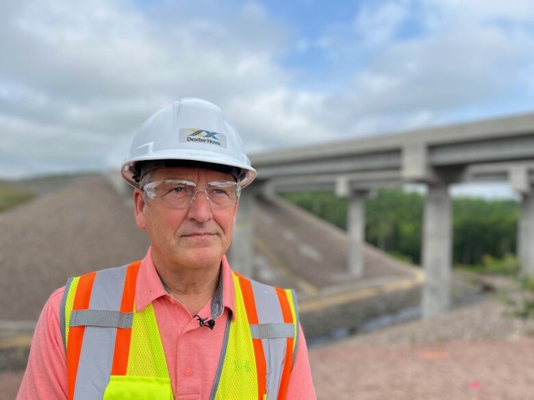 A man with a hard hat and glasses stands in front of a bridge.