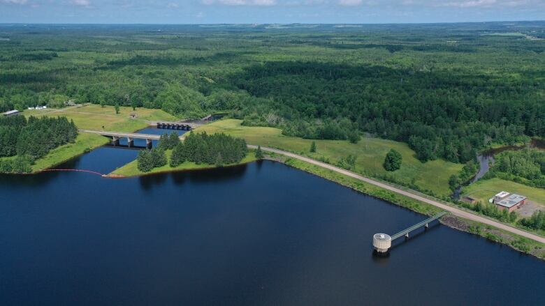 An aerial view of a large lake surrounded by green vegetation with a dam and concrete spillway structure on the top left. A round concrete intake structure is on the right.