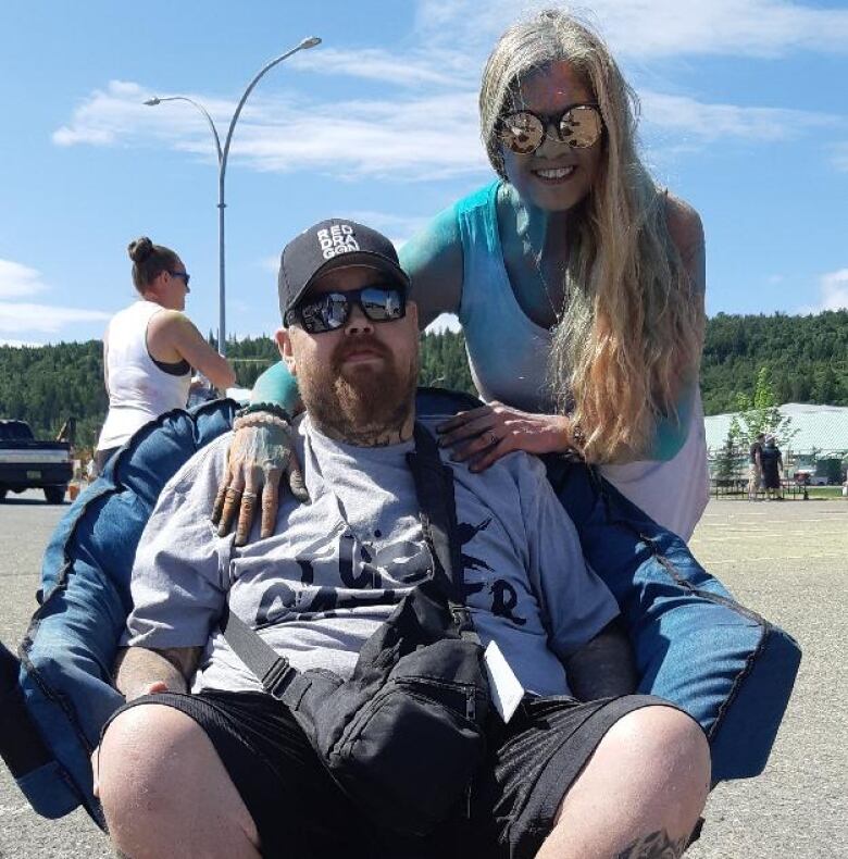 A woman stands behind a man who is seated outdoors in a folding chair.