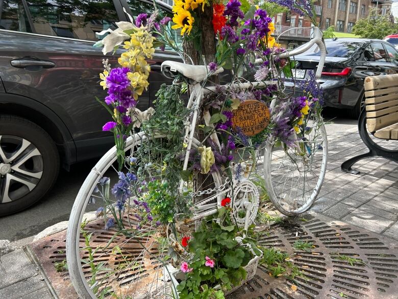 A bike painted white attached to a tree is embellished with flowers. A sign reads 