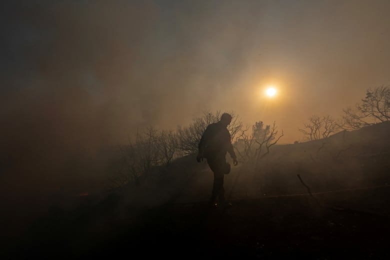 A man walks along a densely smoky hillside near trees, as the sun shines through the smoke.