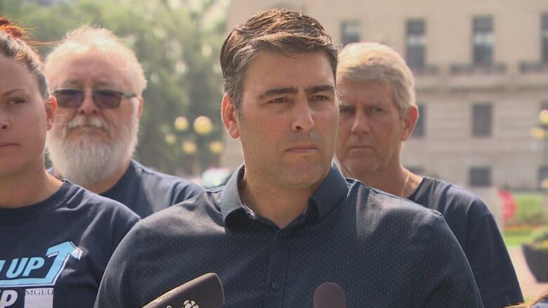 A man with short, black hair, parted to the side, speaks at a podium. He has other people standing beside and behind him.