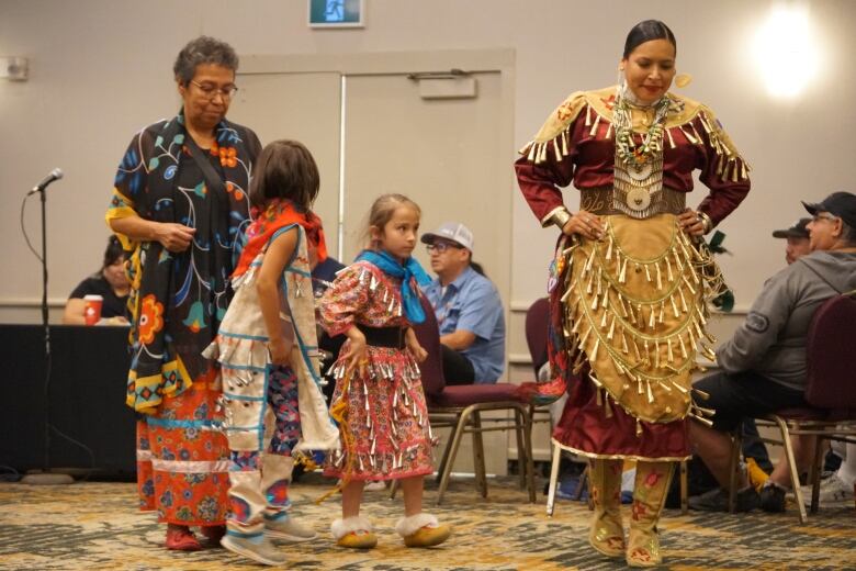 Women and girls dance in traditional Indigenous regalia at a powwow inside a conference room.