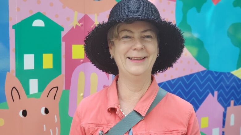 A woman in a coral jean jacket and a black straw sun hat smiles in front of a brightly coloured van.