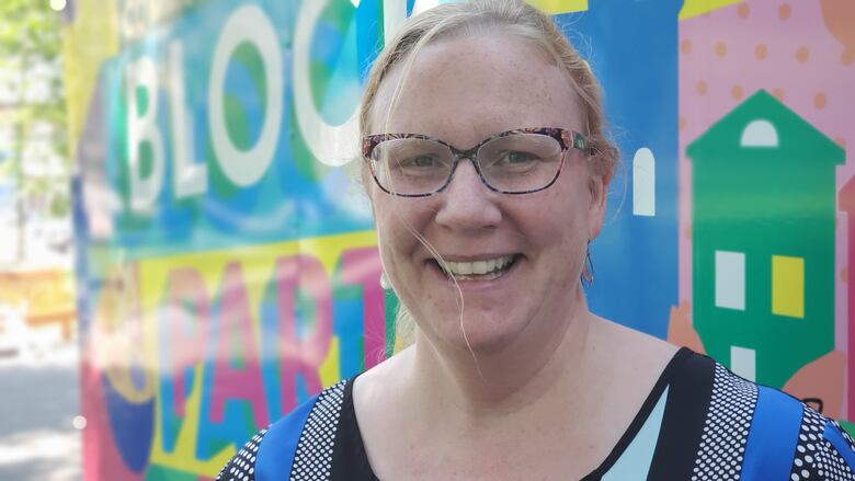 A woman wearing glasses with blonde hair up in a bun stands smiling in front of a colorful van. 