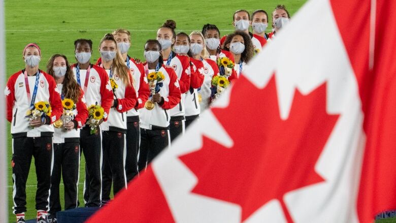 A Canadian flag waves in the forefront as soccer players stand on the podium in the background.