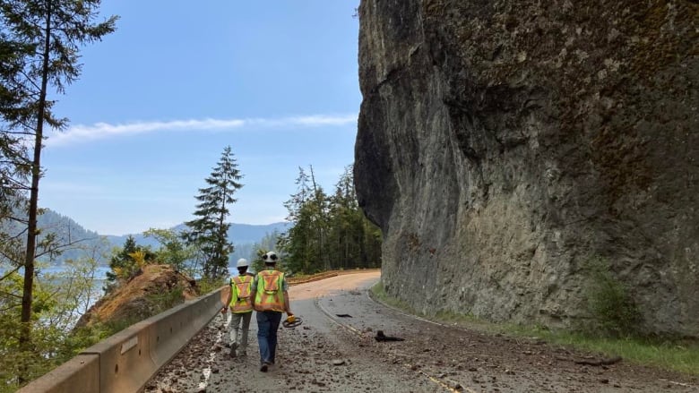 Two people wearing high-vis vests walk along a highway.