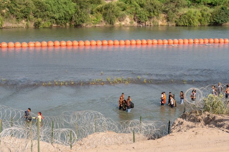 Several people are shown standing in a river from a view on high. In the background, a long orange string of connected buoys is shown.