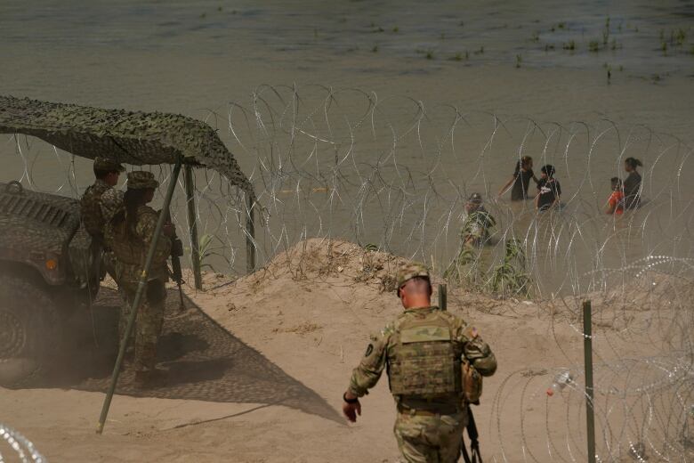 People in military fatigues are shown near a river bank that has razor wire set up.