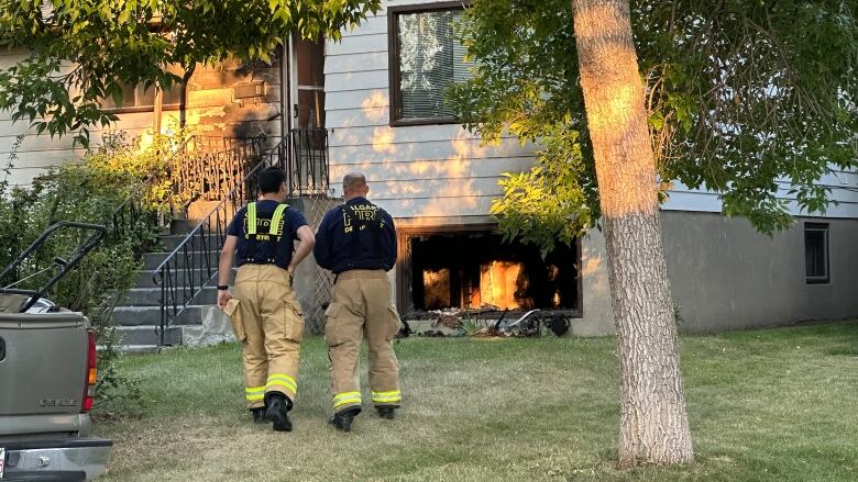 two firemen seen from behind approach a fire damaged house on the front lawn beside a tree.