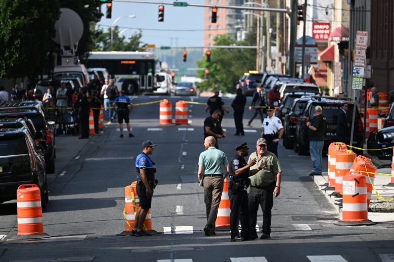 Several people in police uniforms stand on a city street closed off by orange pylons.