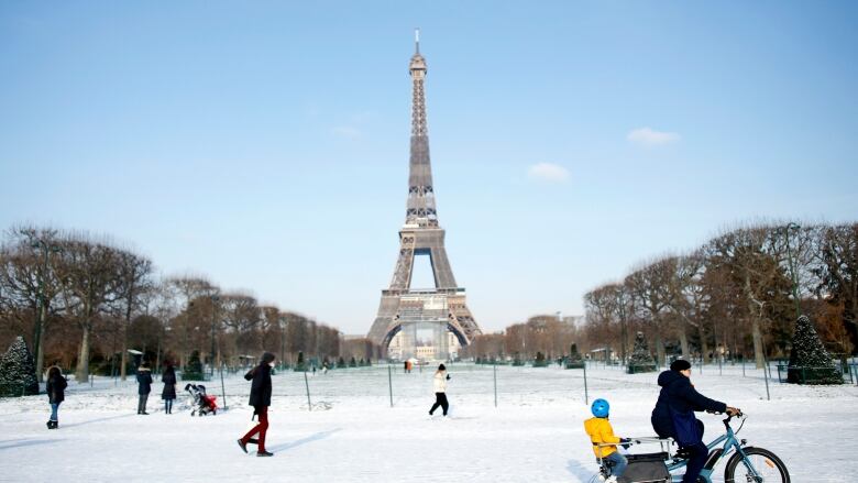 Eiffel Tower in background with people in the foreground, including a man on a bicycle with a child at the back of it.