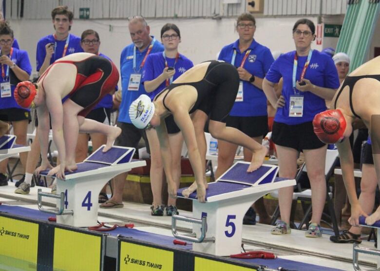 A young woman is poised on the edge of a pool ready to dive in. She wears a black bathing suit and a white swim cap.