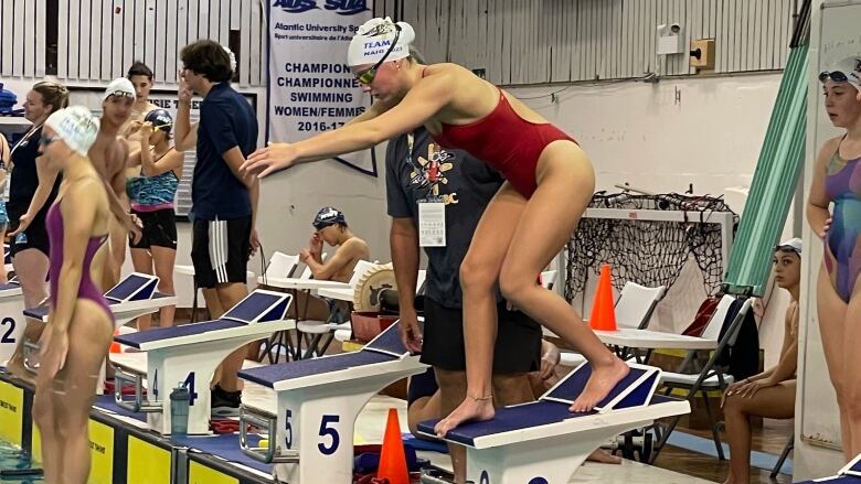 A young woman stands poised to dive into a pool. She wears a red bathing suit and a white swim cap.