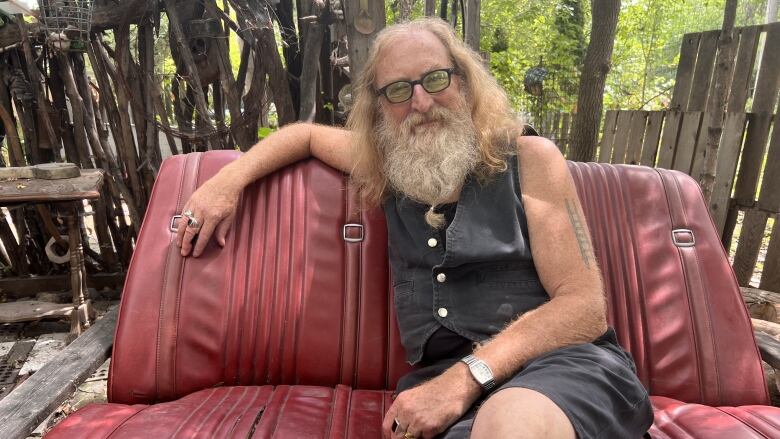 A man is sitting outside on a red couch. A wooden fence is behind him.