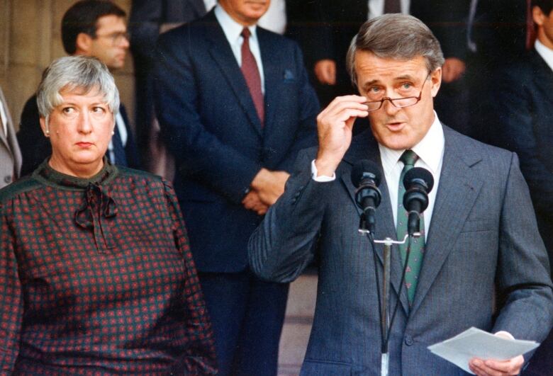 A white-haired woman with short hair stands next to a white man adjusting his glasses in front of a mic.