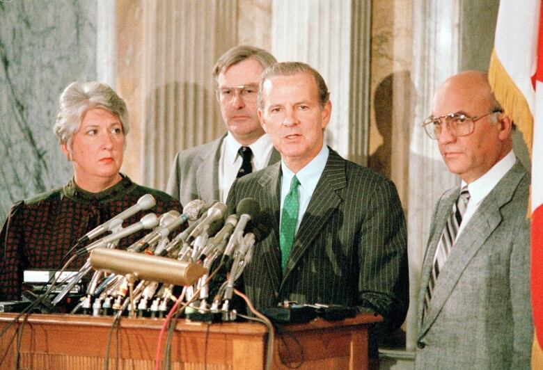 A woman, left, looks on as a man, flanked by two other men, speaks in front of a bank of microphones.