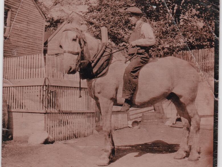 Sepia toned photo of a man sitting on a tall horse in front of a wooden fence. 