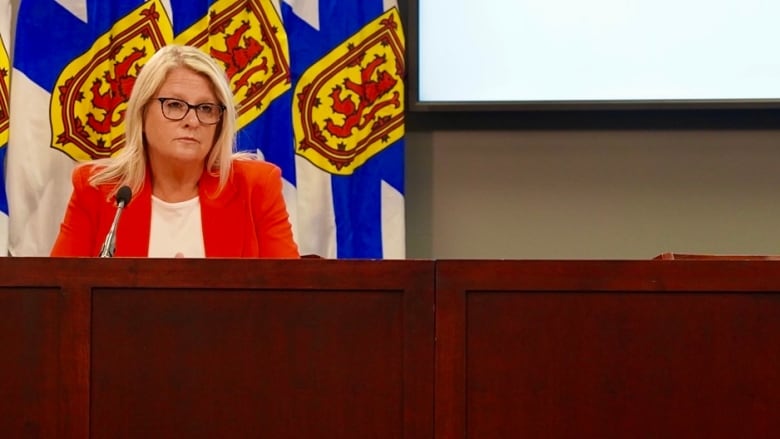 A woman with glasses sits at a podium with flags behind her.