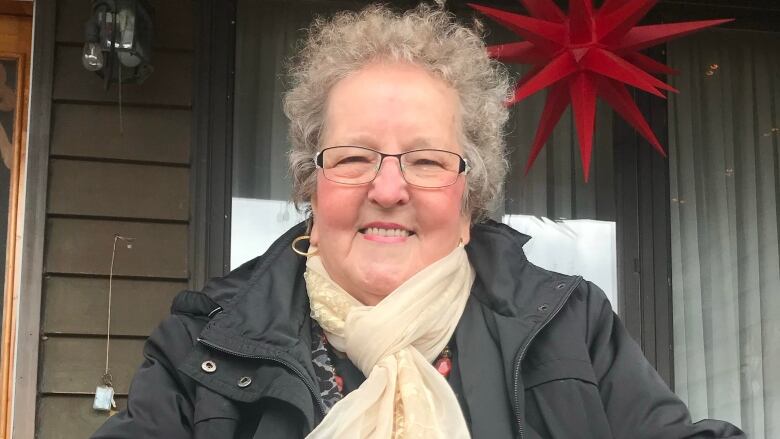 An elderly woman is smiling at the camera. She wears a cream-coloured scarf, black glasses, a black jacket, gold hoop earrings and has curly greyish hair. 