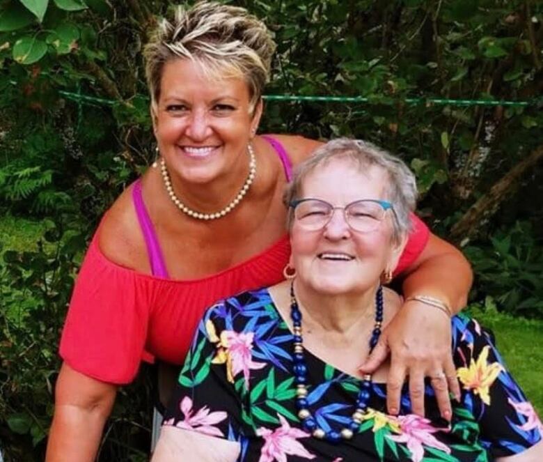 A woman has her arm around an elderly woman. The younger woman has short blonde hair and wears a bright pink top. The elderly woman wears glasses and a top with blue, yellow and pink flowers. 